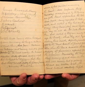 A pair of hands holds open a visibly aged journal, showing handwritten text inside. The journal appears small