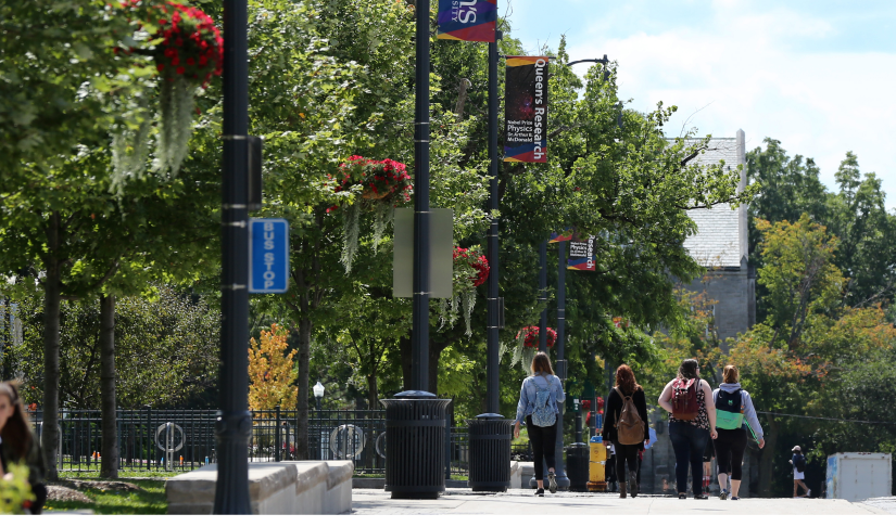 Students crossing the street on campus. Lots of leafy trees and "Queen's Research" banners hanging from lamp posts.