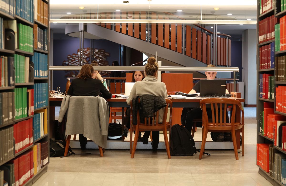 Four students sitting at a table on their laptops surrounded by library book shelves