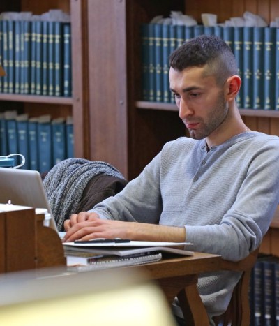 Student working on his laptop with books in the background. 