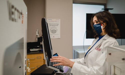 A woman wearing a mask and lab coat types at a computer